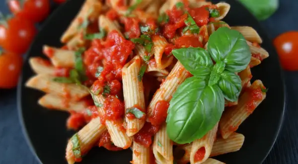 Penne z sosem pomidorowym i świeżą bazylią, ozdobione listkami bazylii. Obok pomidorki koktajlowe.