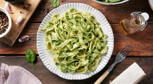 Pyszne dania z makaronem tagliatelle z zielonym pesto, podane na talerzu z niebieskimi paskami. Obok świeży szpinak, czosnek i przyprawy.
