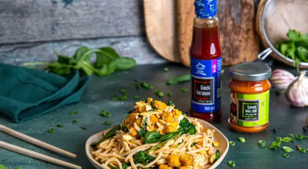 Pyszny makaron po koreańsku z tofu, szpinakiem i sosem kimchi. Obok butelka sosu i słoiczek pasty czosnek-imbir-chili.