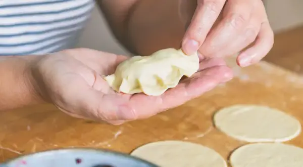 Ręce formują pieroga według przepisu. Na blacie leżą okrągłe placki ciasta i miska z wiśniami.