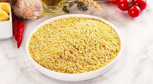 Miseczka pełna drobnego makaronu orzo, obok czosnek, papryczki chili, pomidorki i oliwa.
