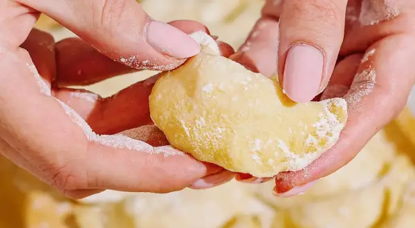 Dłonie z różowymi paznokciami formują ciasto do pierogów, obsypane mąką. W tle widać gotowe pierogi.