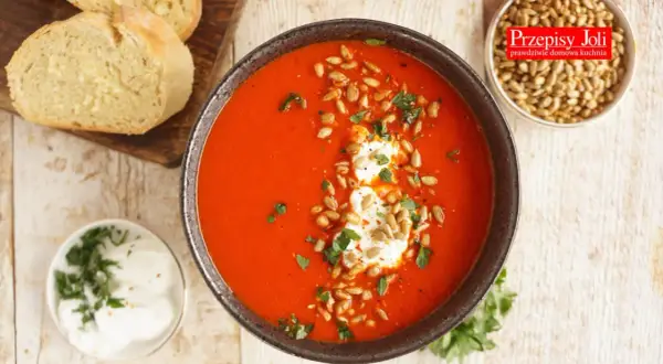 Aksamitny krem z pomidorów i papryki, udekorowany śmietaną, pestkami słonecznika i natką pietruszki. Obok pieczywo i miseczka z dodatkami.