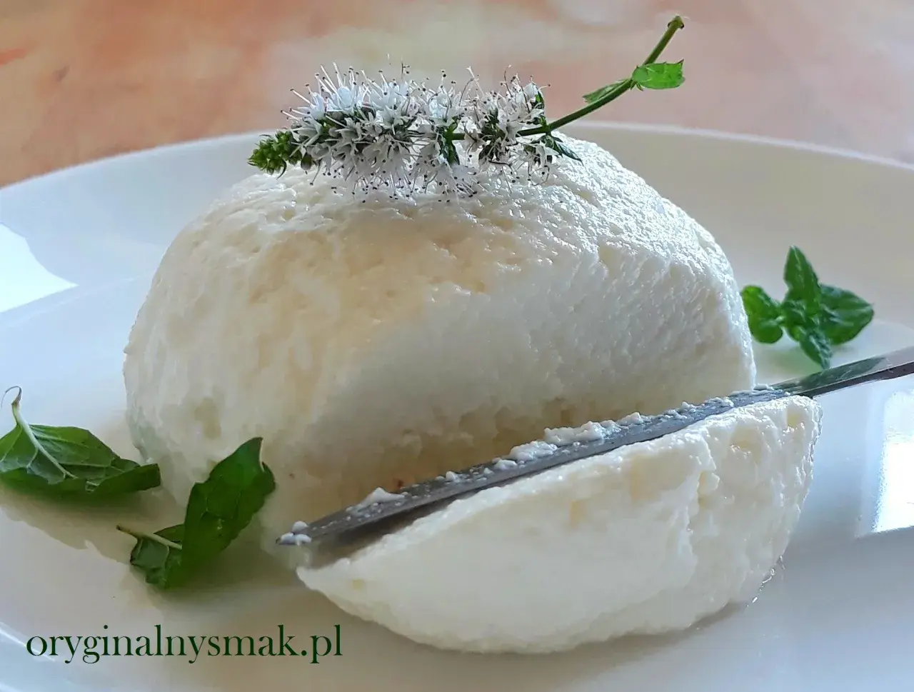 Puszysta, domowa ricotta na talerzu, ozdobiona kwiatami mięty. Nóż delikatnie kroi ser, odsłaniając jego kremową konsystencję.