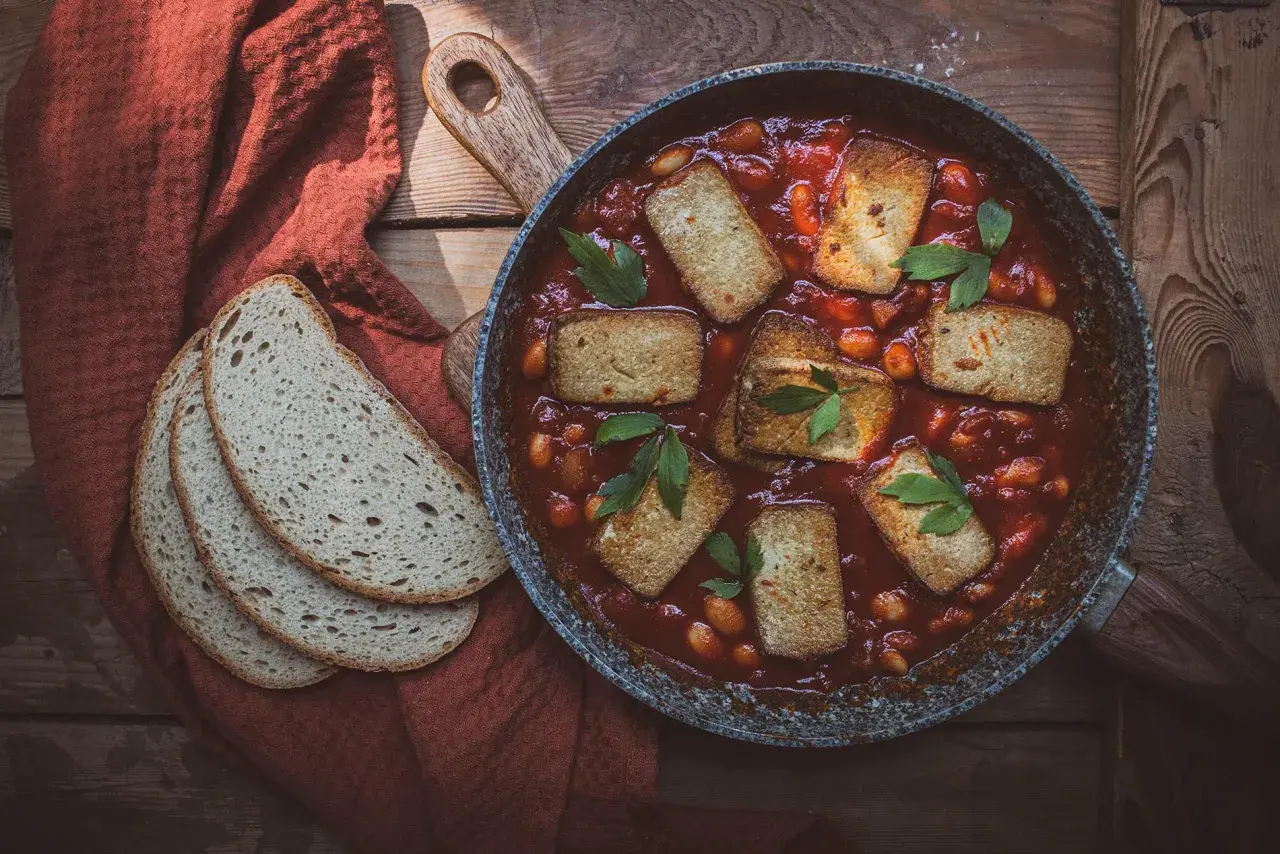 Aksamitne tofu w sosie pomidorowym z fasolką, podane w patelni, obok kromek chleba na rustykalnym stole.