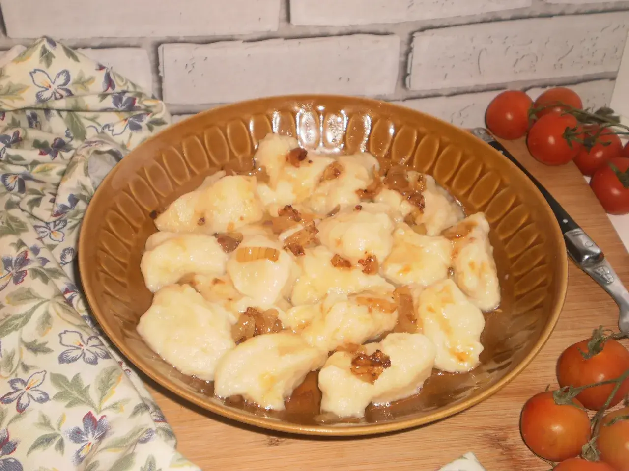 Pyszne kopytka ruskie polane skwarkami z cebulki, podane na brązowym talerzu, obok gałązka pomidorków koktajlowych.