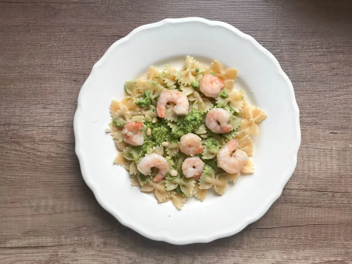 Makaron farfalle z zielonym pesto, krewetkami i orzeszkami pinii na białym talerzu.
