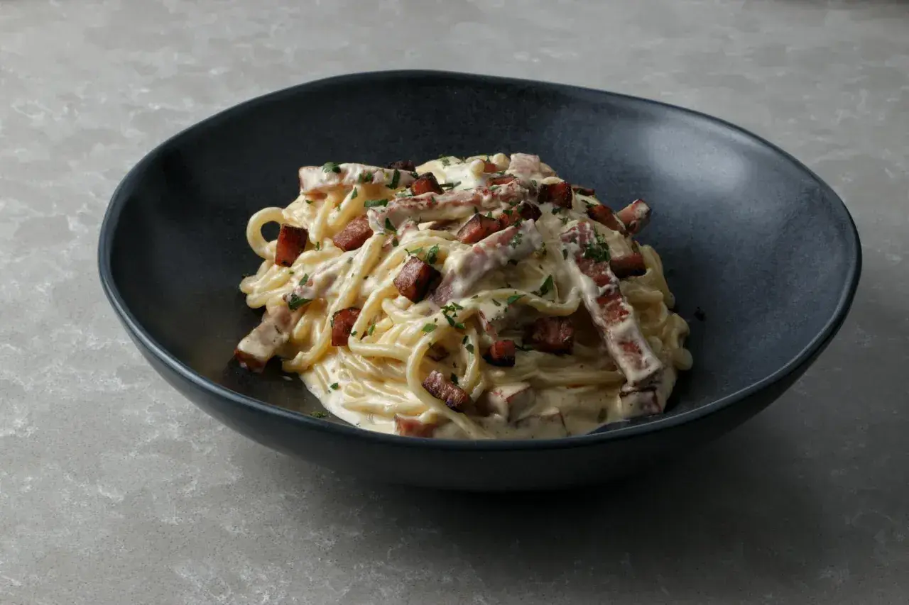 Pyszny przepis na carbonare: spaghetti z kremowym sosem, boczkiem i natką pietruszki w ciemnej misce.