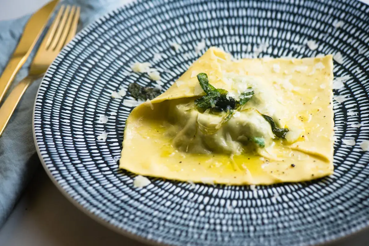 Duży raviolo con uovo, polany masłem i posypany parmezanem, na talerzu z czarno-białym wzorem.