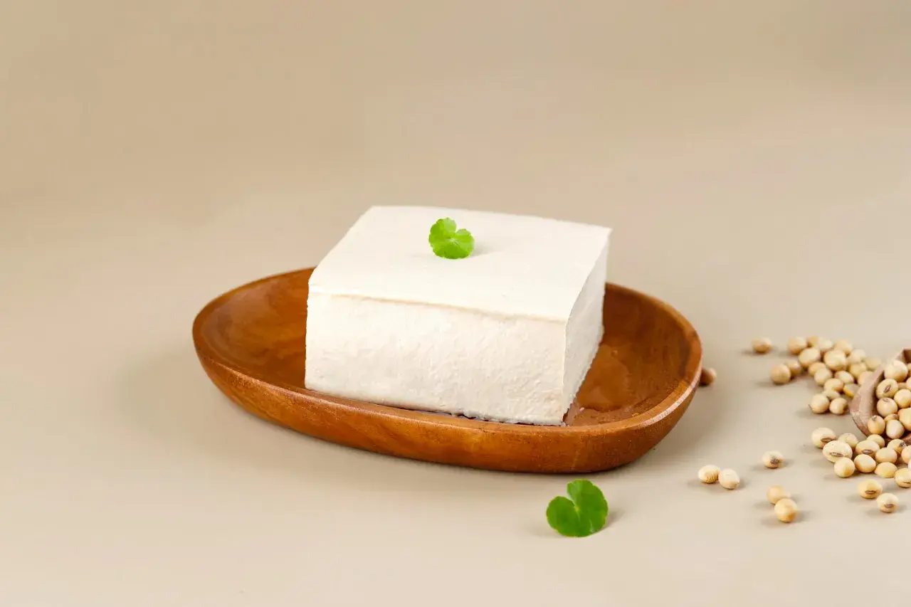 Kostka białego tofu na drewnianej desce, obok nasiona soi. Różne rodzaje tofu są wszechstronne w kuchni.