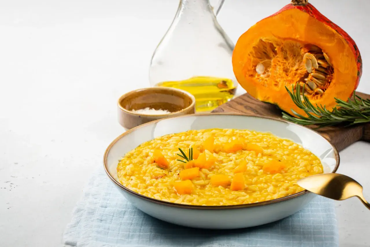 Kremowe risotto dyniowe w misce, z kawałkami dyni i rozmarynem. Obok połówka dyni, oliwa i sól.