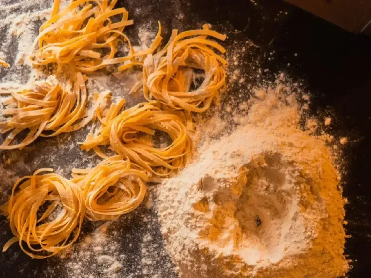 Świeże, domowe tagliatelle uformowane w kółka, leżą obok kopczyka mąki na ciemnym blacie.