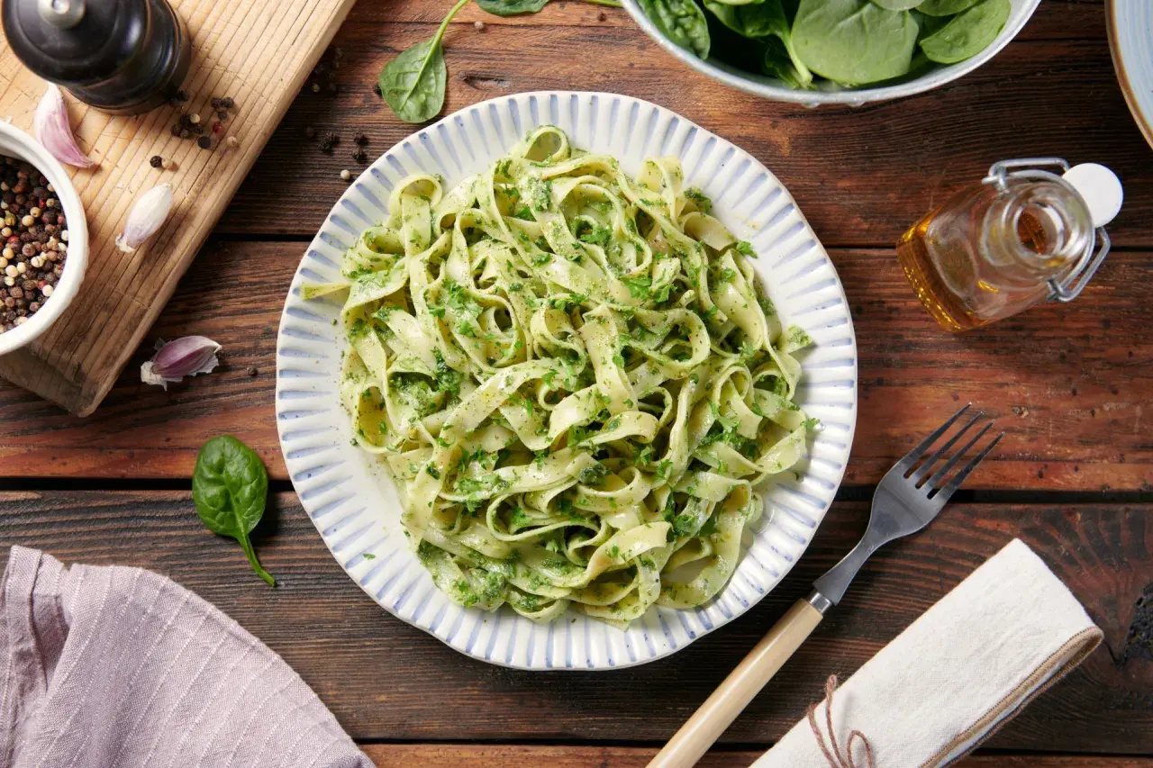 Pyszne dania z makaronem tagliatelle z zielonym pesto, podane na talerzu z niebieskimi paskami. Obok świeży szpinak, czosnek i przyprawy.