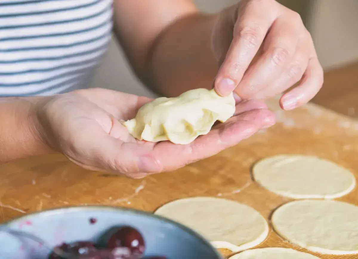 Ręce formują pieroga według przepisu. Na blacie leżą okrągłe placki ciasta i miska z wiśniami.