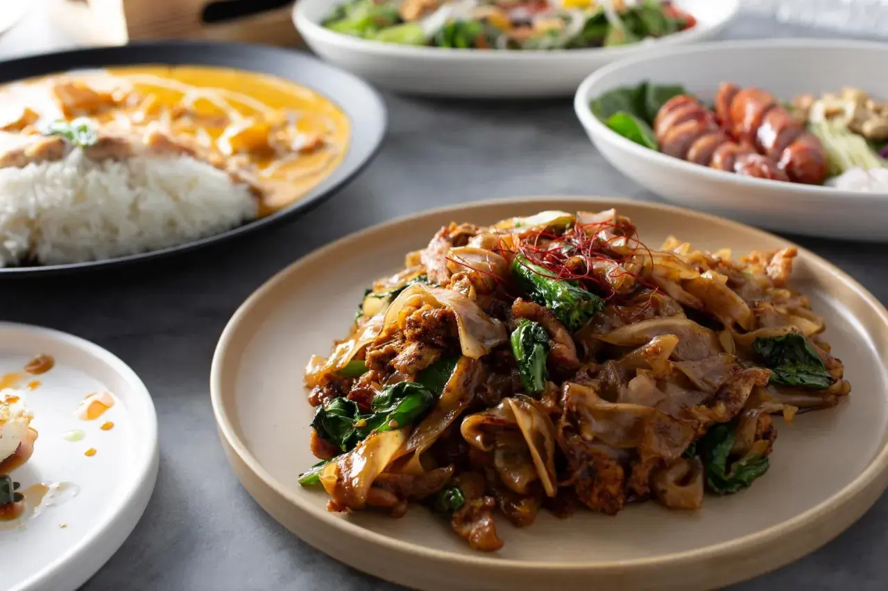 Pyszny pad thai makaron z kurczakiem i warzywami, podany na talerzu. Obok widać inne dania.