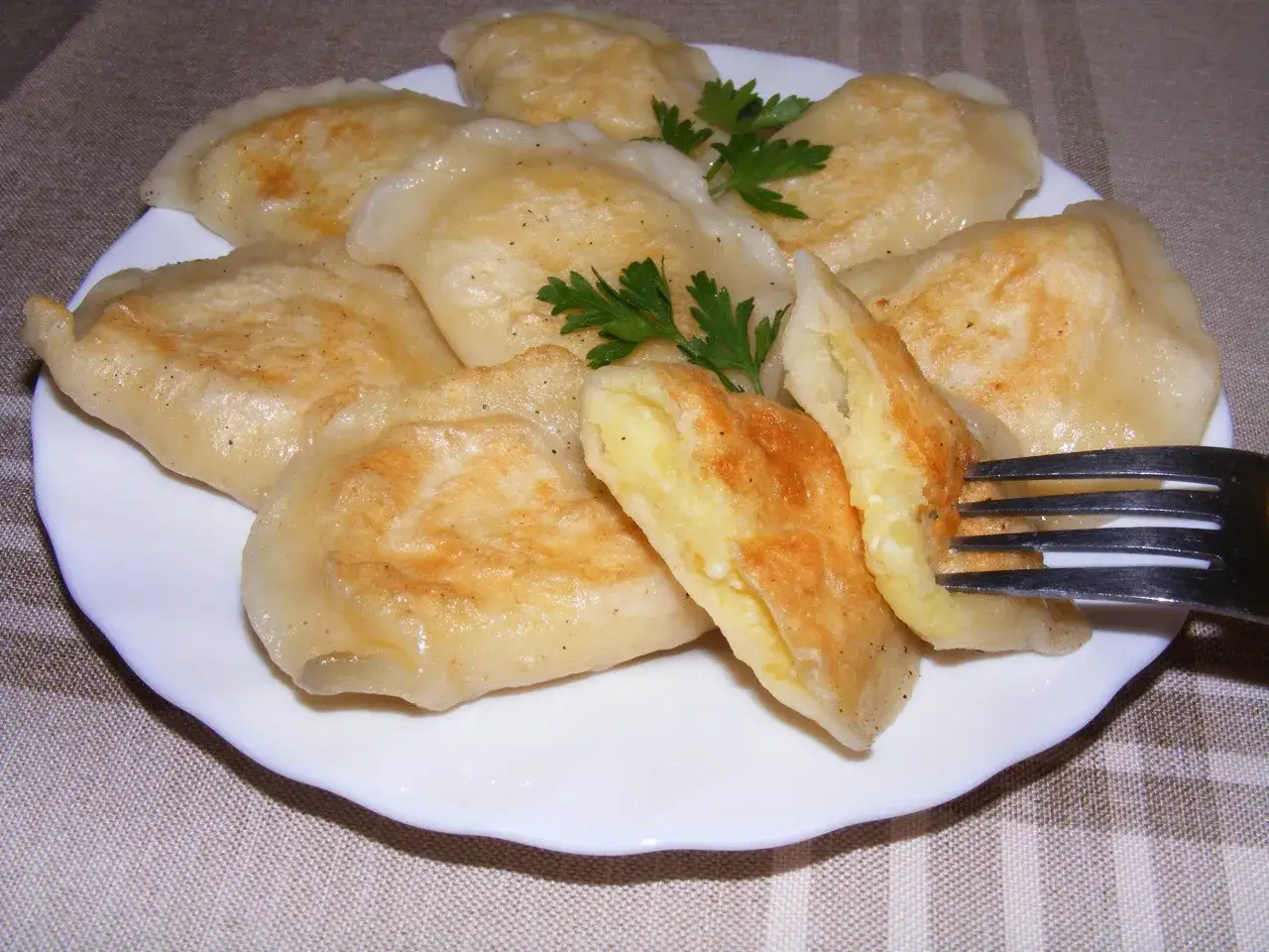 Talerz pełen pysznych, złocistych pierogów z ziemniakami, jeden z nich jest nadziany na widelec, ozdobiony gałązką natki pietruszki.