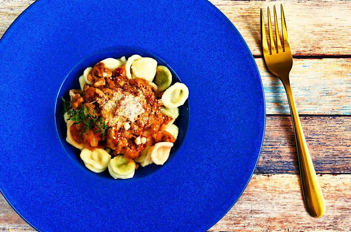 Pyszne makaron orecchiette z sosem mięsnym i parmezanem na niebieskim talerzu, obok złoty widelec.