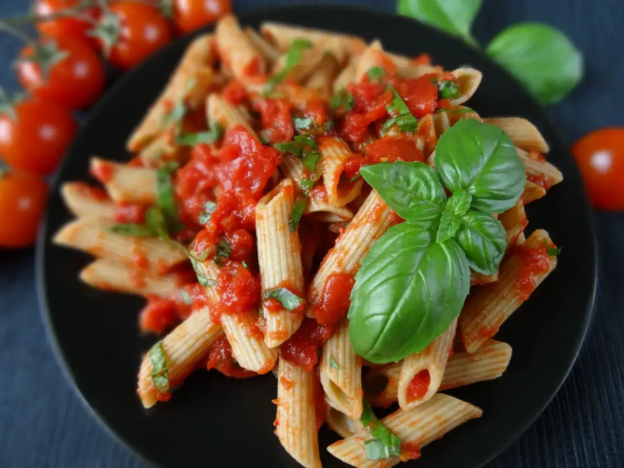 Penne z sosem pomidorowym i świeżą bazylią, ozdobione listkami bazylii. Obok wiązka pomidorków koktajlowych.
