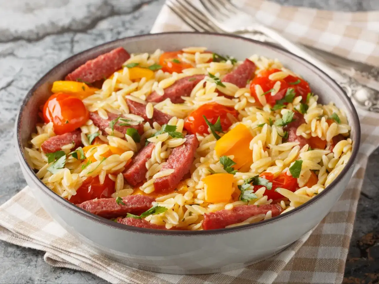 Pyszne danie z makaronem orzo, kawałkami pikantnej kiełbasy, pomidorkami koktajlowymi i papryką, posypane świeżą natką pietruszki.