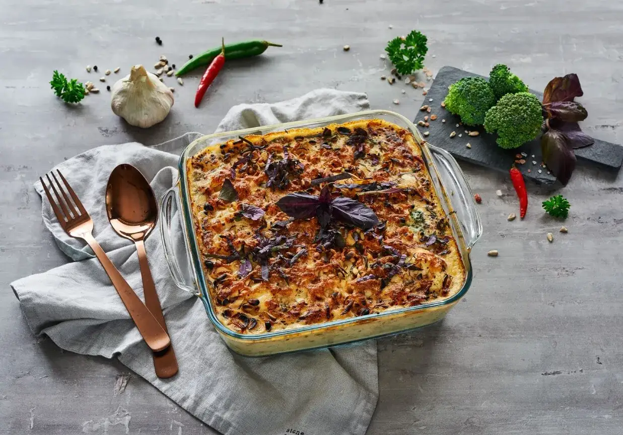 Pyszna lasagne wegetariańska w szklanym naczyniu, ozdobiona bazylią. Obok sztućce, czosnek, papryczki chili i brokuły.
