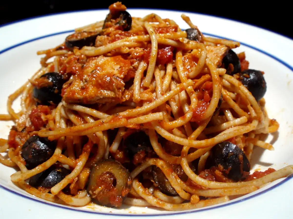 Spaghetti z tuńczykiem po włosku, z kawałkami ryby, pomidorowym sosem i czarnymi oliwkami, podane na białym talerzu.