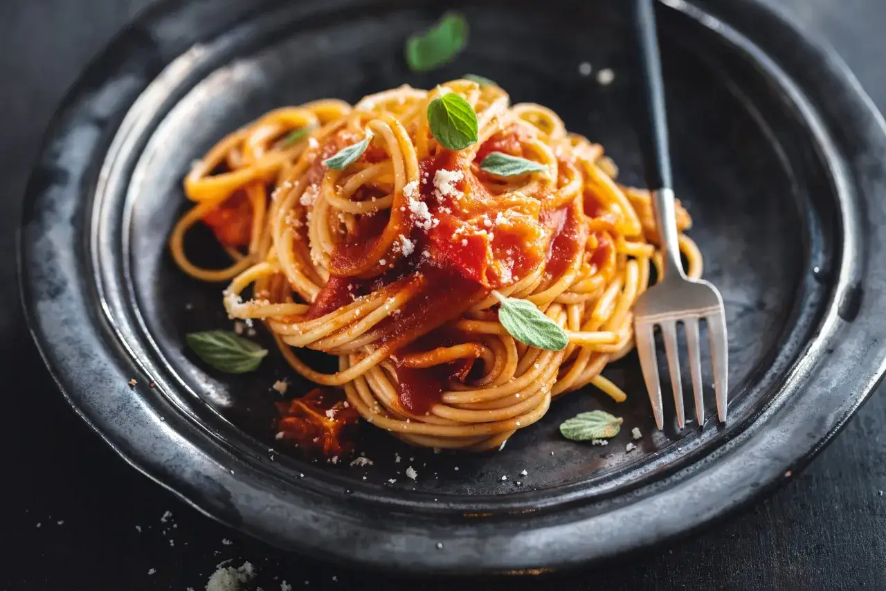 Pyszna włoska pasta spaghetti z sosem pomidorowym, posypana parmezanem i udekorowana listkami bazylii. Obok leży widelec.