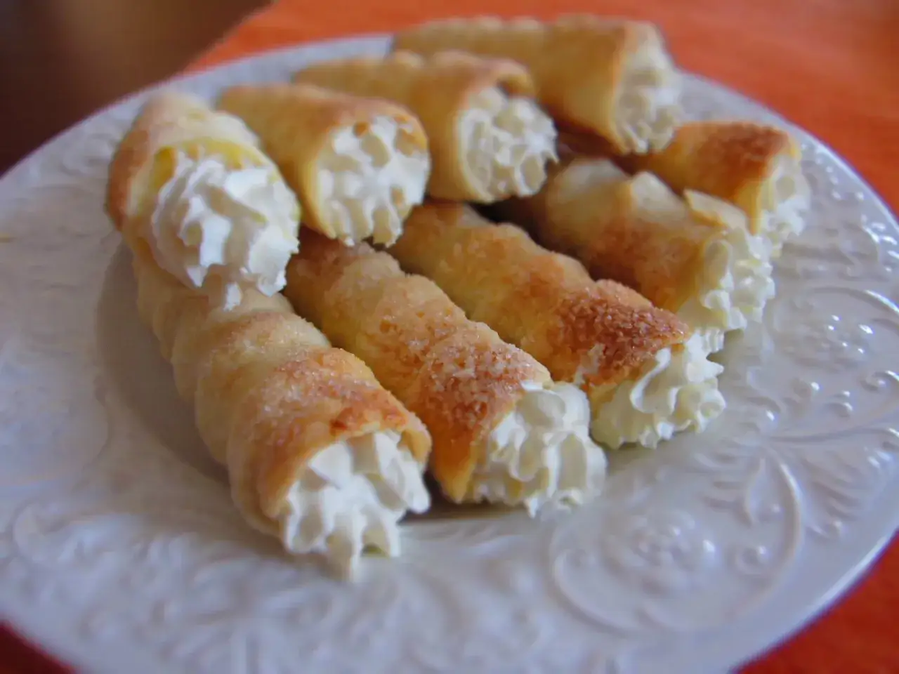 Chrupiące rurki z kremem do cannoli, posypane cukrem, ułożone na białym talerzu.