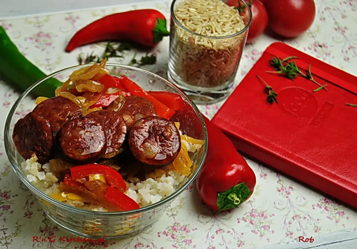 Danie z chorizo, ryżem i papryką w misce. Obok szklanka ryżu, czerwona papryka i czerwony notes.