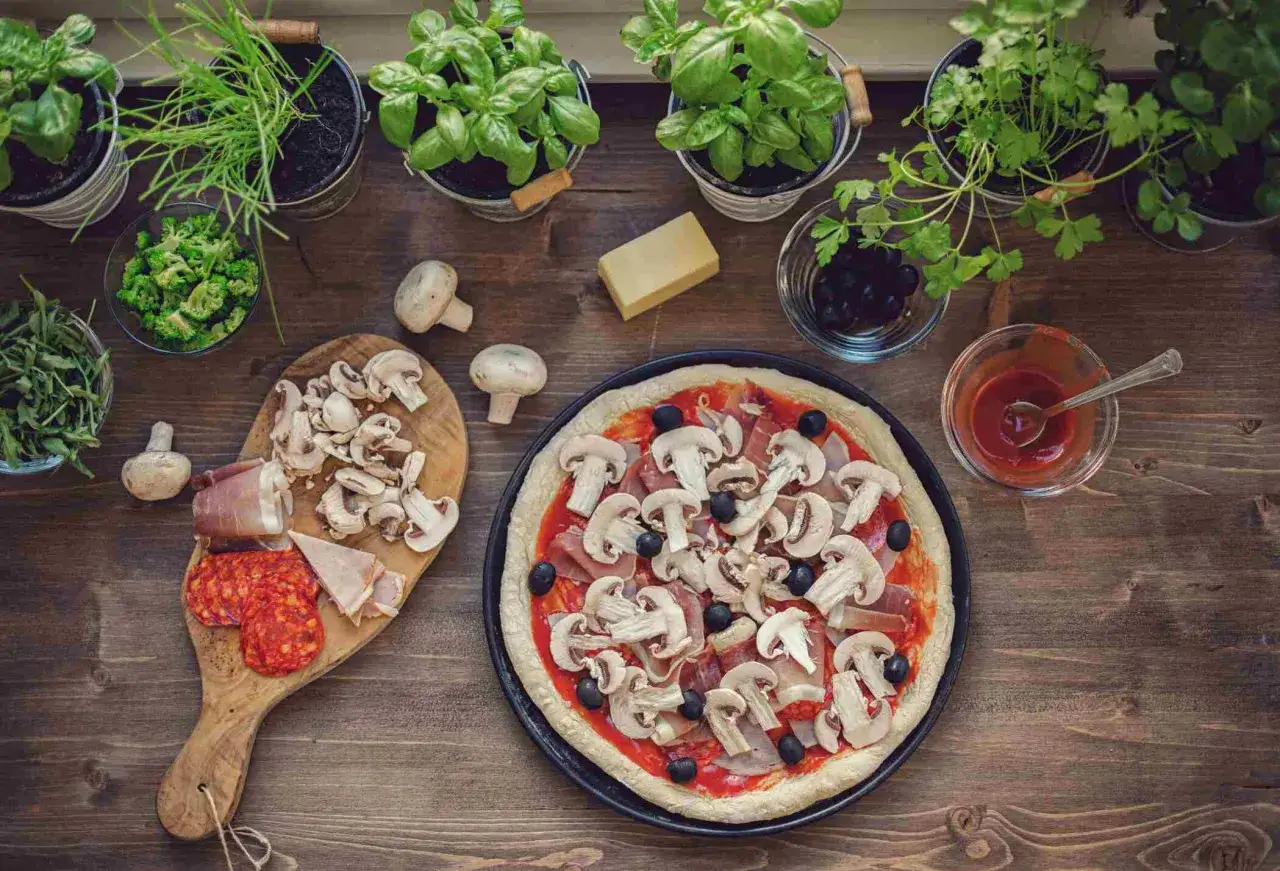 Przygotowania do pizzy: świeże zioła, pieczarki, oliwki, salami i szynka jako dodatki do pizzy.
