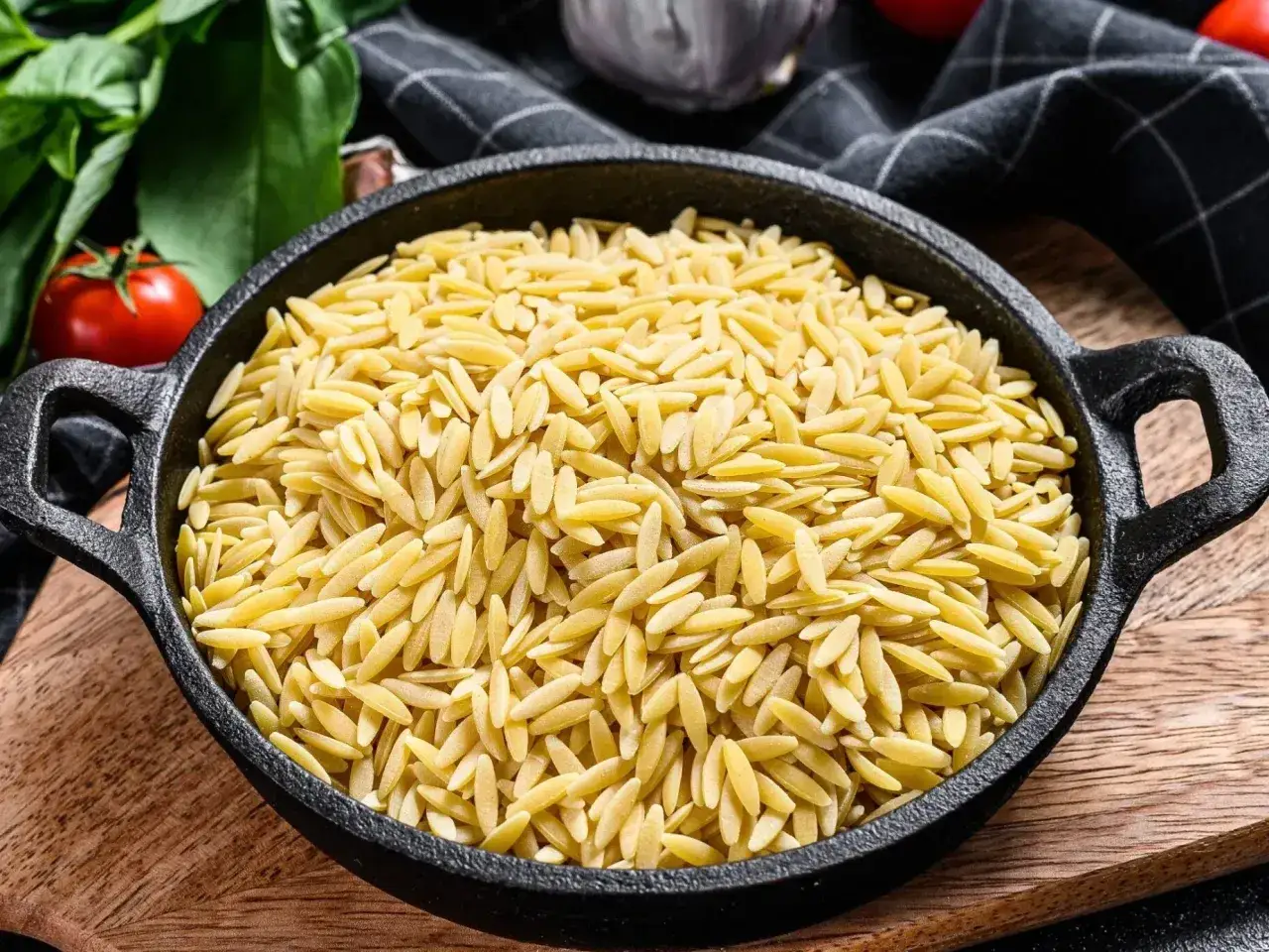 Makaron orzo w czarnej patelni, gotowy na pyszny przepis. Obok pomidory i zielenina.