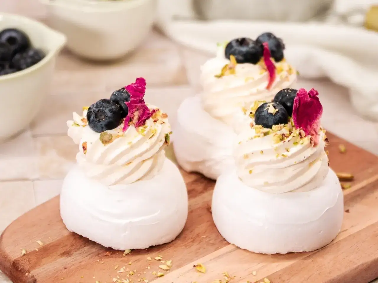 Mini bezy pavlova z bitą śmietaną, borówkami i płatkami róży. Pyszne, delikatne deserki na drewnianej desce.