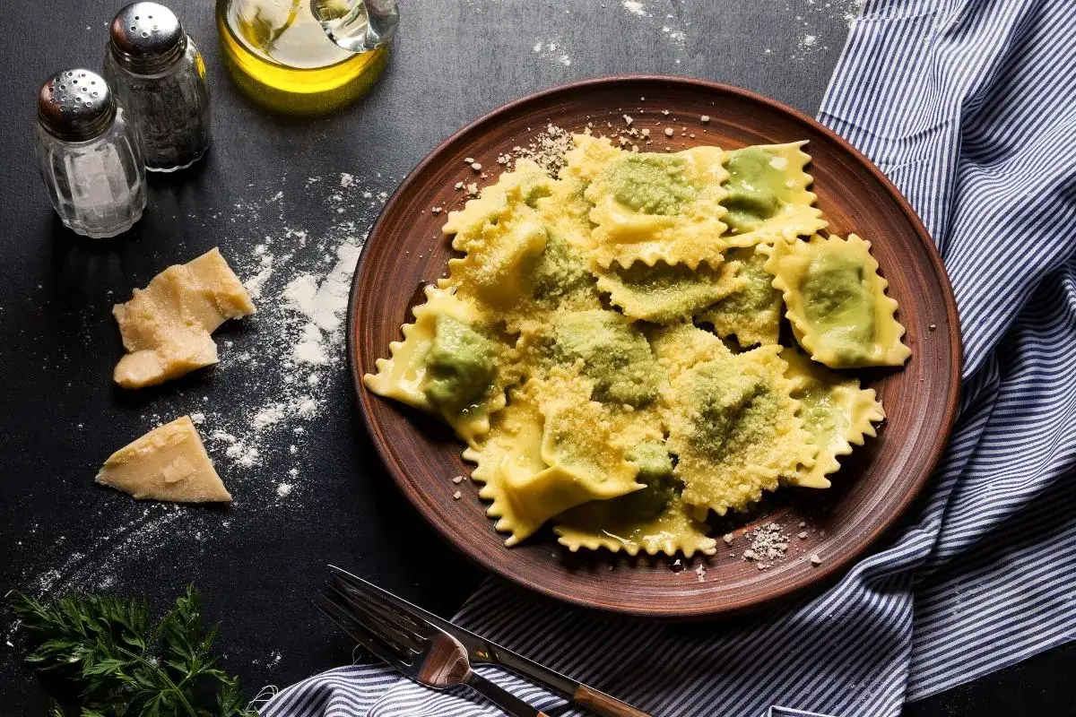 Pyszne ravioli ze szpinakiem i ricottą, posypane parmezanem, na brązowym talerzu. Obok kawałki sera i oliwa.
