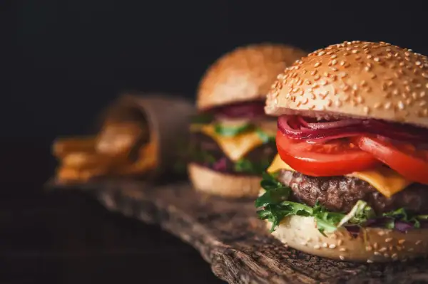 Dwa soczyste smash burgery z serem, pomidorem i czerwoną cebulą, obok papierowego rożka z frytkami.