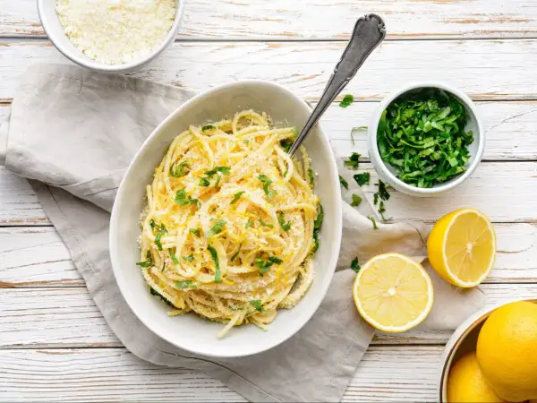 Pyszny makaron cytrynowy posypany parmezanem i świeżą natką pietruszki, obok połówki cytryny.