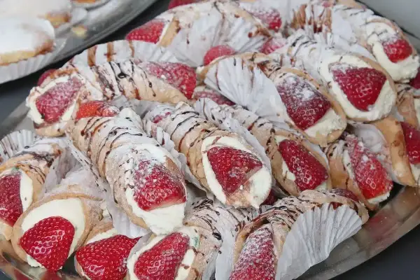 Taca pełna pysznych cannoli z kremem i truskawkami, posypana cukrem pudrem i polana czekoladą.