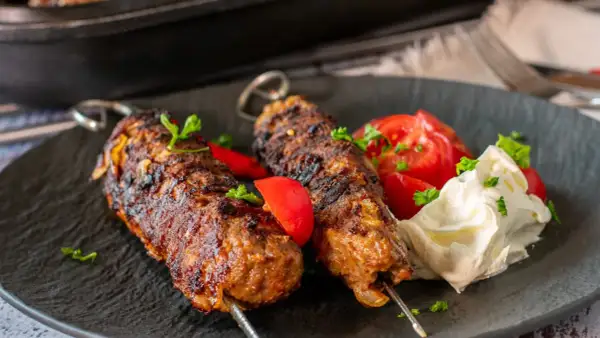 Dwa soczyste kofty na grilla, podane z pomidorami i kremowym dipem, ozdobione natką pietruszki. Idealne na letnie przyjęcie.