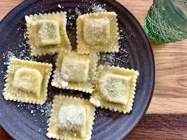 Pięć kwadratowych ravioli z ricottą, posypanych parmezanem, na ciemnym talerzu. Obok zielony kieliszek.