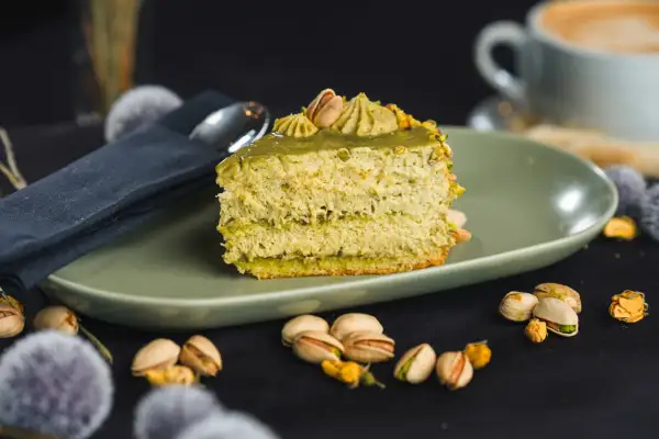 Pyszny kawałek tortu pistacjowego, udekorowany kremem i orzechami. Obok filiżanka kawy i łuskane pistacje.
