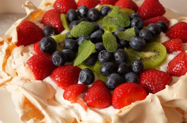 Pyszny tort bezowy z owocami: truskawki, borówki, kiwi i listki mięty na kremie.