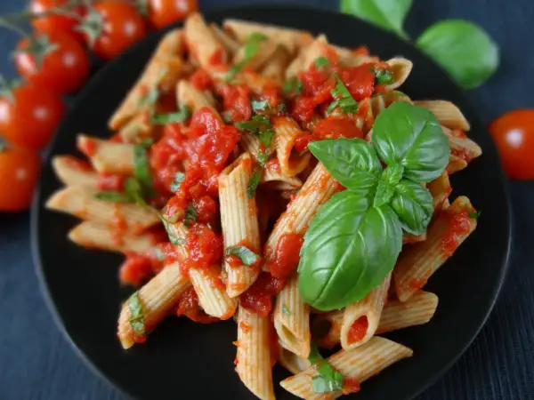 Penne z sosem pomidorowym i świeżą bazylią, ozdobione listkami bazylii. Obok pomidorki koktajlowe.