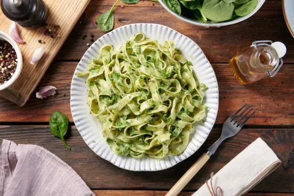 Pyszne dania z makaronem tagliatelle z zielonym pesto, podane na talerzu z niebieskimi paskami. Obok świeży szpinak, czosnek i przyprawy.