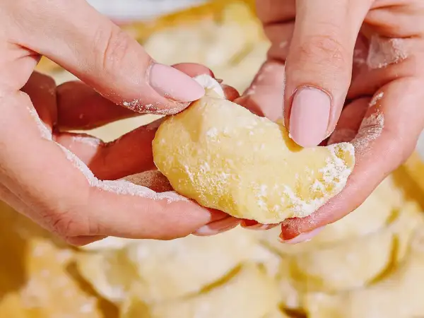 Dłonie z różowymi paznokciami formują ciasto do pierogów, obsypane mąką. W tle widać gotowe pierogi.
